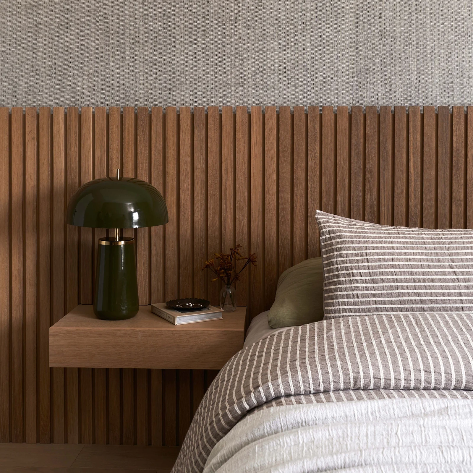 Interior Design - A bedside table with a green lamp, books, and a vase on a wooden surface next to a bed with striped bedding. Dexign Matter Studio