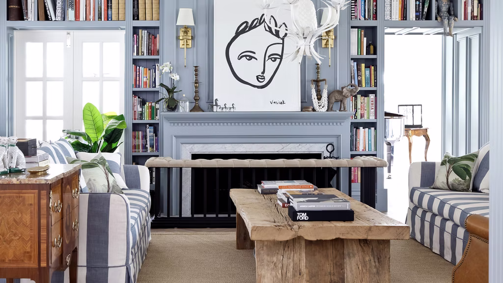 Stylish living room with blue shelves, striped sofas, a wooden table, and a fireplace. Artwork of a face above. Bright and cozy ambiance.