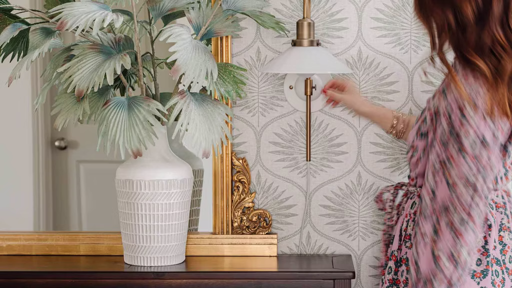 Green wallpaper. A wall sconce beside a mirror on a console table. A vase with leaves sits on a dark wooden table.