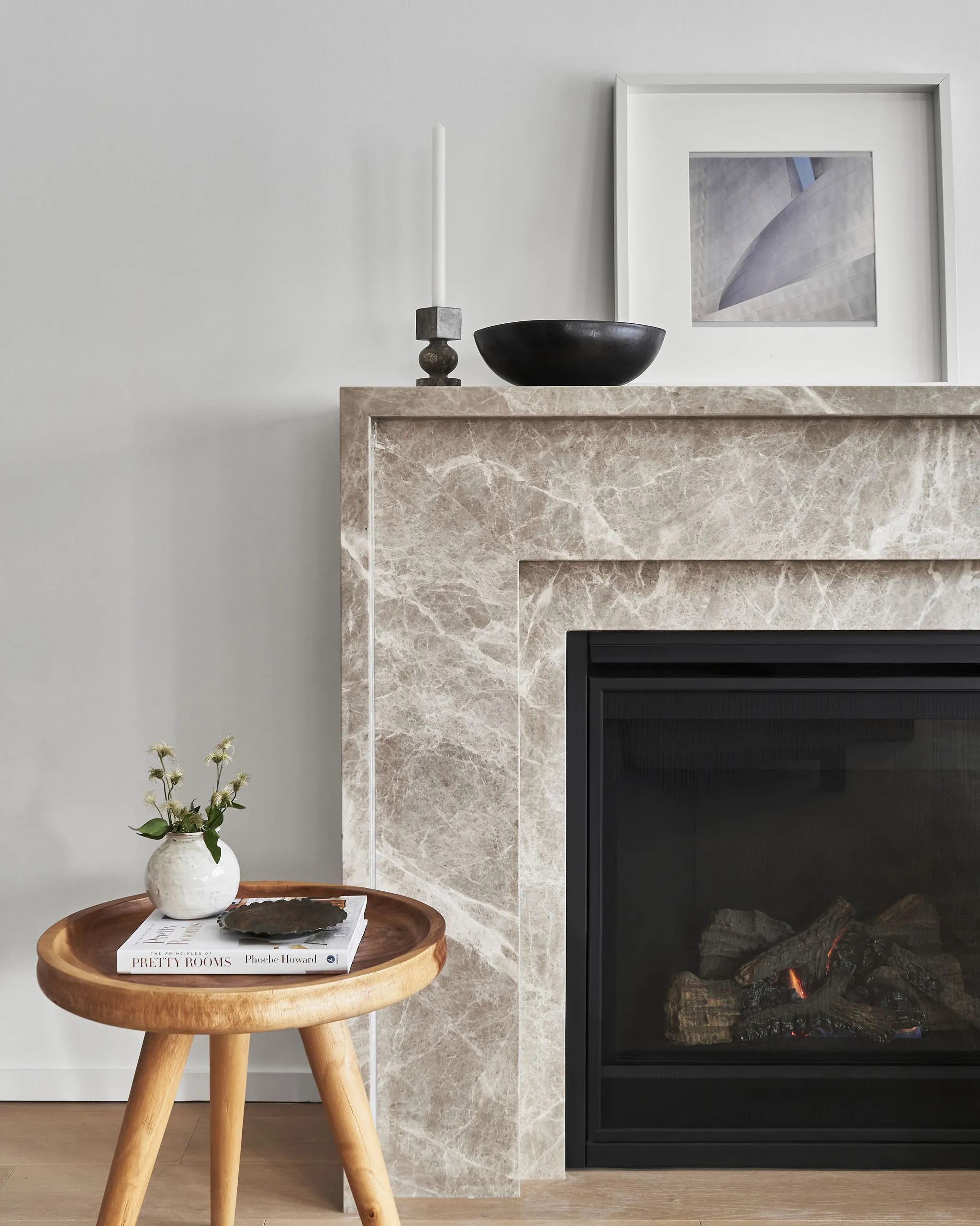 Interior Design - A marble fireplace featuring a black bowl, candle holder, and a wooden side table with a vase and books. Dexign Matter Studio